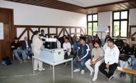 Mehrere Gäste sind von vorne zu sehen und schauen zur Rednerin, deren Rücken zu erkennen ist. Sie steht neben einem Tisch mit Projektor.