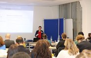 Eine Frau steht vor einer Gruppe sitzender Zuhörer*innen. Im Hintergrund wird eine Präsentation gezeigt.