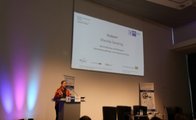 Einige Gäste sind von hinten zu sehen und schauen nach vorne zur Rednerin, die vor einer Präsentation steht.
