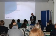 Ein Mann steht vor einer Gruppe sitzender Personen. Im Hintergrund wird eine Präsentation gezeigt.