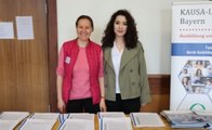 Zwei Frauen stehen an einem Tisch mit Infoblättern. Neben ihnen ist das Plakat der KAUSA-Landesstelle Bayern zu erkennen.