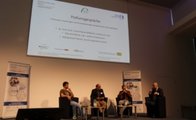 Vier Personen sitzen in Sesseln auf dem Podium. Im Hintergrund ist eine Präsentation zu sehen auf der "Podiumsgespräch" steht.