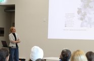 Die Moderatorin des Keynote-Vortrag blickt zur Präsentation. Von einigen Gästen sind die Hinterköpfe zu sehen.