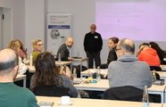 Ein Mann steht vor einer Gruppe diskutierender Gäste. Im Hintergrund wird eine Präsentation gezeigt.