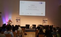 Mehrere Gäste sind von hinten zu sehen und schauen nach vorne zur Rednerin, die vor einer Präsentation steht.