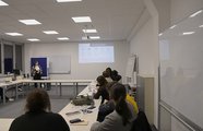 Eine Frau steht vor einer Gruppe sitzender Zuhörer*innen. Im Hintergrund wird eine Präsentation gezeigt.