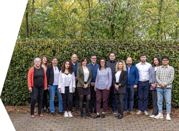 KAUSA-Team in Bayern Mitarbeiter*innen der KAUSA-Landesstelle Bayern stehen als Gruppe vor einer grünen Hecke.
