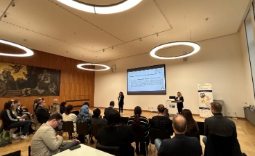 Mehrere Gäste sitzen an ihren Tischen und schauen nach vorne zu den beiden Rednerinnen vor einer Präsentation.