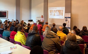 Mehrere Gäste sind von hinten zu sehen und schauen nach vorne zum Redner, der vor einer Präsentation steht.