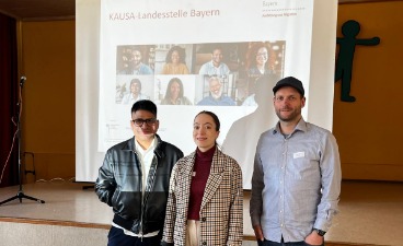 Drei Personen stehen vor einer Präsentation der KAUSA-Landesstelle Bayern.