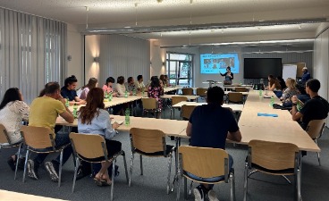 Gäste der Informationsveranstaltung sitzen in einem großen Raum an Tischen, die in U-Form angeordnet sind. Sie sind überwiegend von hinten zu sehen und schauen zu einer Präsentation nach vorne.