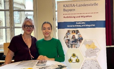 Zwei Frauen stehen nebeneinander an einem Tisch neben dem Plakat der KAUSA-Landesstelle Bayern