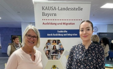Zwei Frauen stehen nebeneinander vor dem Plakat der KAUSA-Landesstelle Bayern