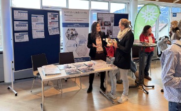Eine Frau spricht an einem Infostand mit einer anderen Frau. Im Hintergrund ist das Plakat der KAUSA-Landesstelle zu sehen.